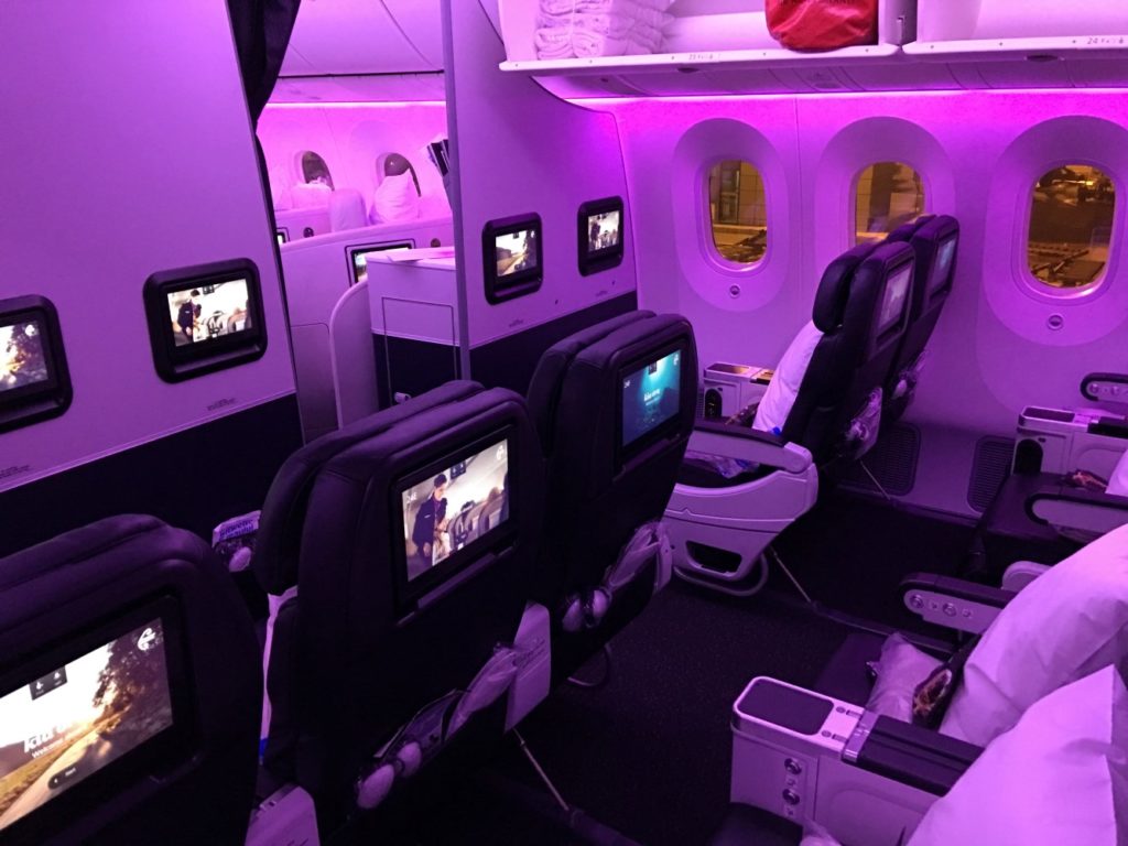 Air New Zealand Premium Economy class section is bathed in purple lighting. An overhead compartment is open and seat back screens are playing a variety of entertainment.