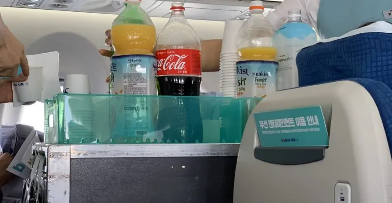 A trolley is wheeled down the aisle with lots of plastic bottles on it. The bottles contain water, soda and juice.