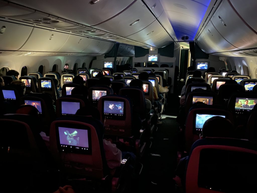 Lights down in the aircraft cabin showing multiple seats and IFE screens displaying different movies