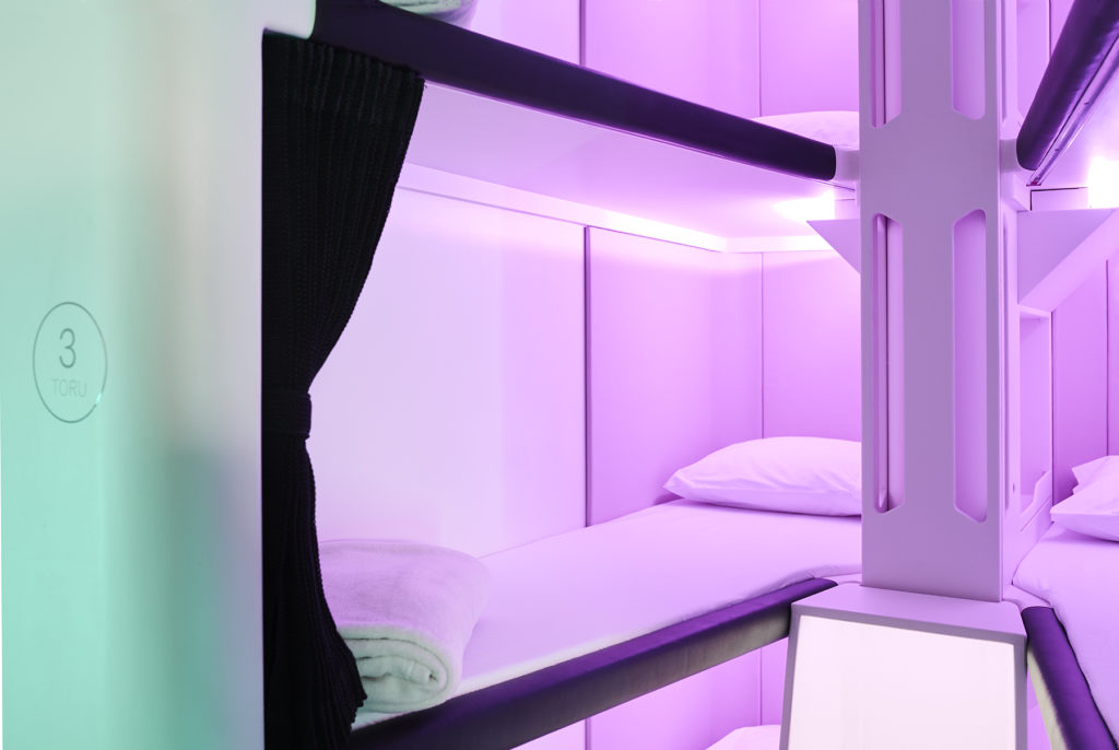 Close up of some of the Skynest bunks with white linens, black curtains, and purprle LED lighting.