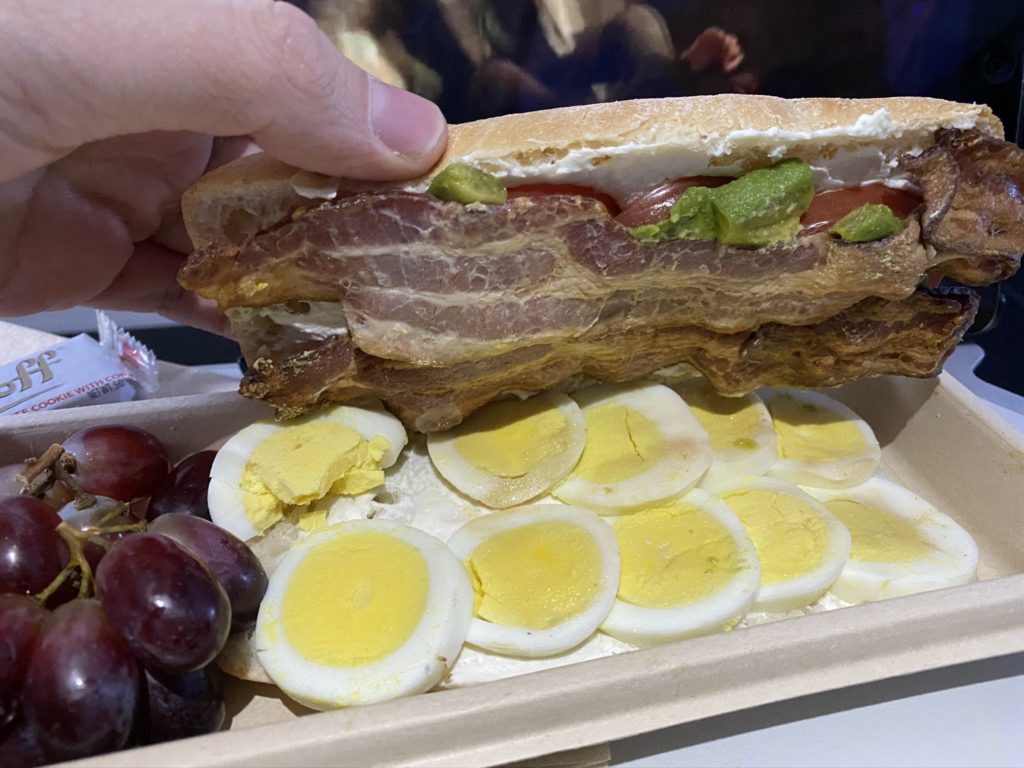 The sunrise breakfast sandwich with someone opening it to see a large layer of sliced hardboiled eggs, layer of bacon and a side of grapes