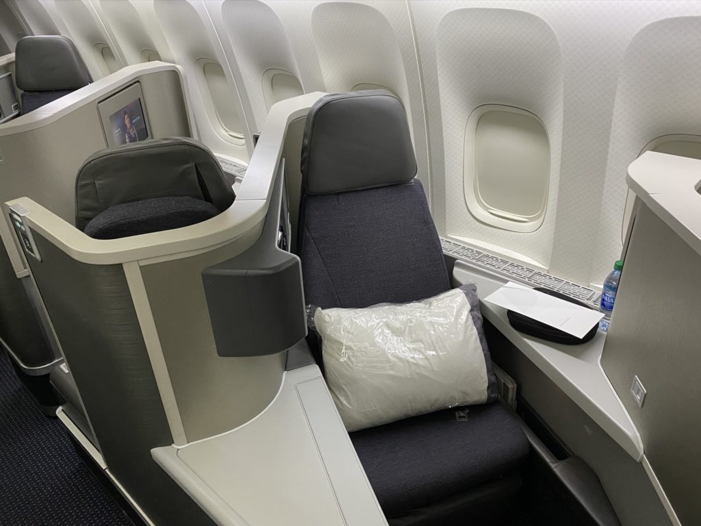 All grey business class seat with a white pillow wrapped in plastic and a menu card with a bottle of water. 