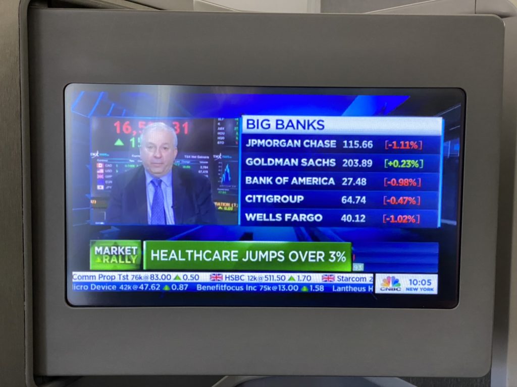 IFE Screen displaying and older man with big banks list and values and a headline that says health care jumps over 3%