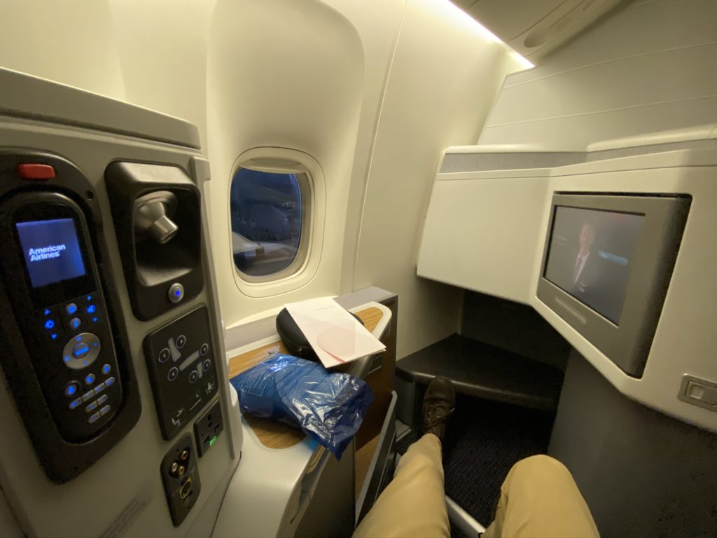 A business class seat, with the remote control and in-seat power sockets to the left and an IFE screen to the right. An aircraft window is also seen in the photo, as well as the author's legs, and a blanket wrapped in plastic