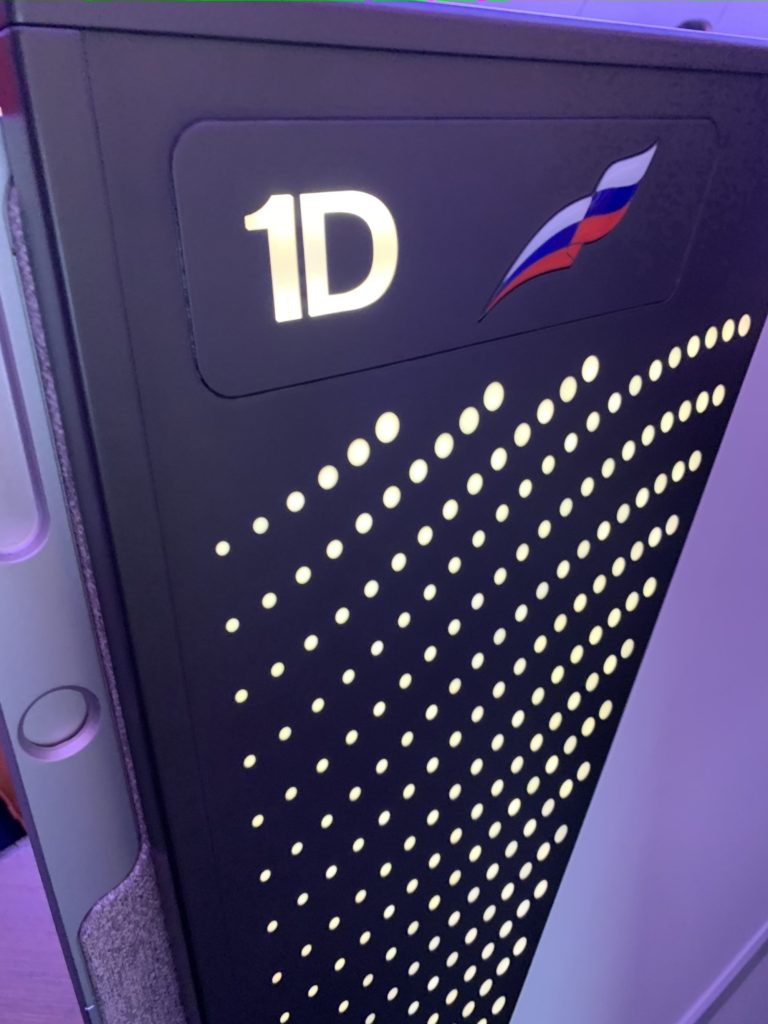 Panel on the business class seat in a dark blue with a white label listing seat "1D" and white dots creating a wave-like image under the Russian flag symbol