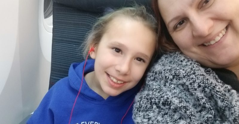 Two passengers, a mother and daughter are smiling for a photo while seated together in an aircraft.