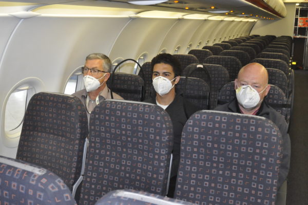 Three men, wearing masks, are seated in a narrowbody aircraft. The image shows the PPW on either side of the middle seat, which protects all passengers