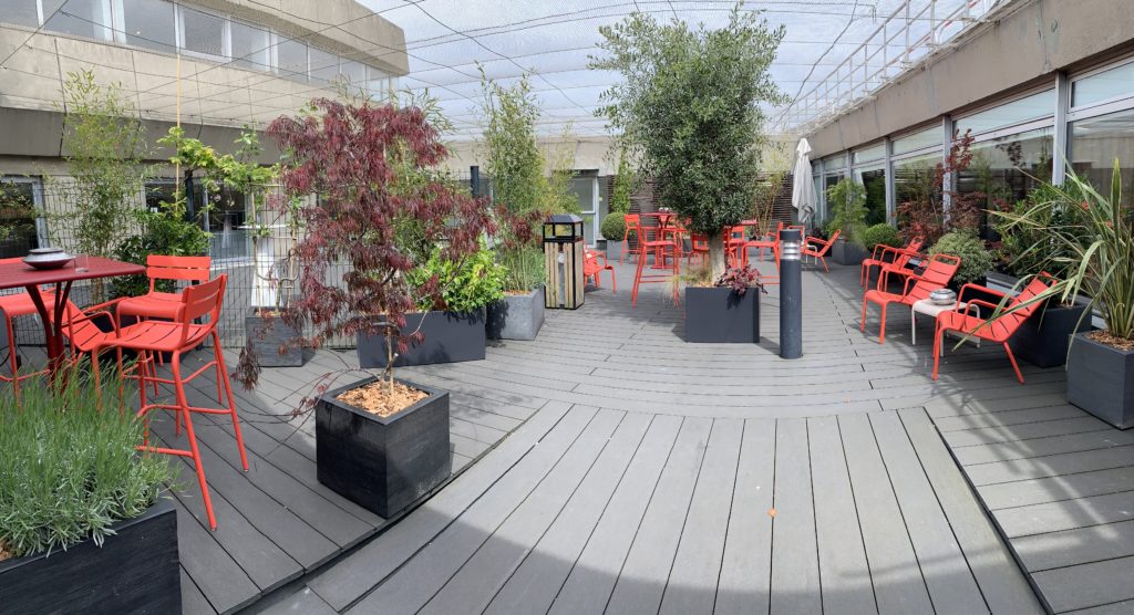 Orange-pink high top tables and chairs, plus regular chairs are seen in the sunshine with a variety of plants.