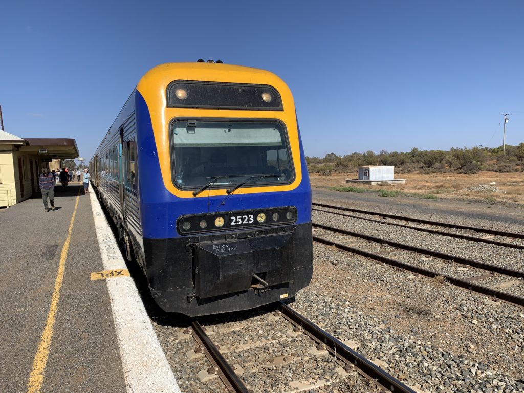 The Outback Xplorer train, in blue and yellow, on the tracks on a beautiful day with blue skies and no clouds