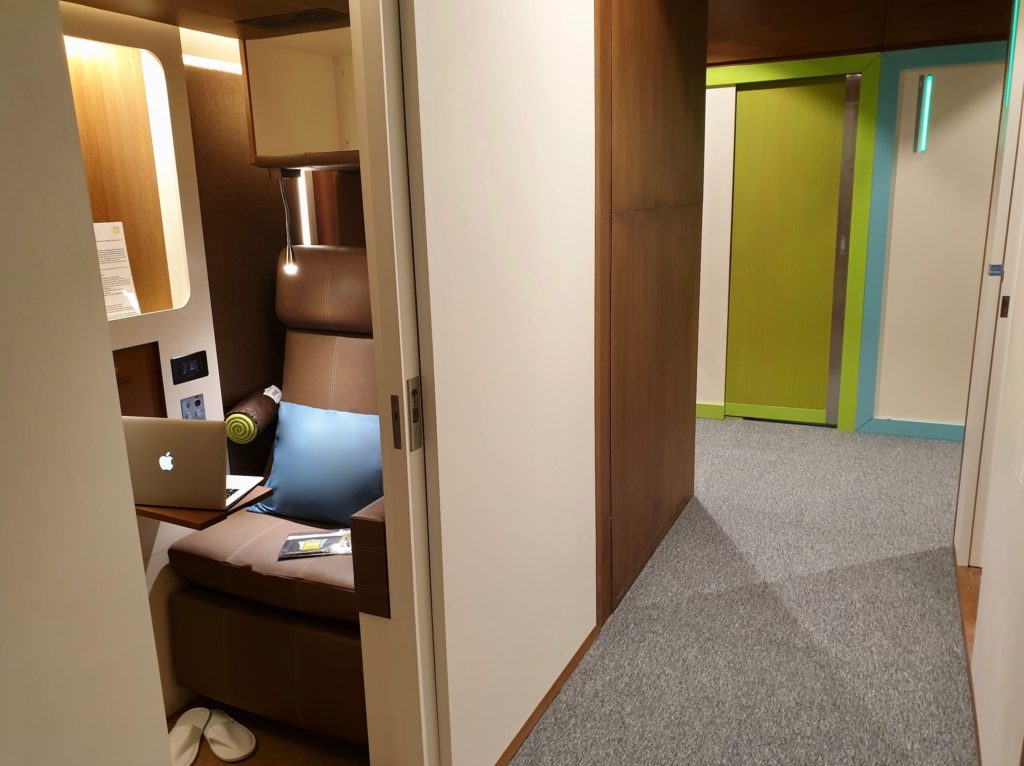 A glimpse into one of the FlexiSuite Pods shows a brown leather chair, and pillow. Slippers are on the floor. The room has soft lighting, and wood accents