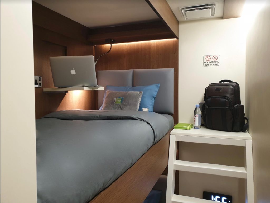 A bunk bed in one of the sleep cabins, with grey bedding and a blue pillow. A bag sits on some shelves in the room