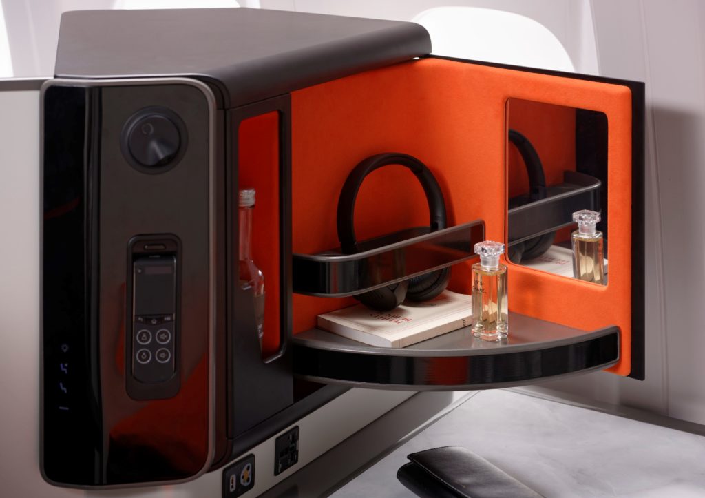 A close-up view of the side storage area of Opera. A headset and a bottle of perfume are displayed in a pull-out compartment. The interior of the compartment is bright orange, and includes a small mirror. 