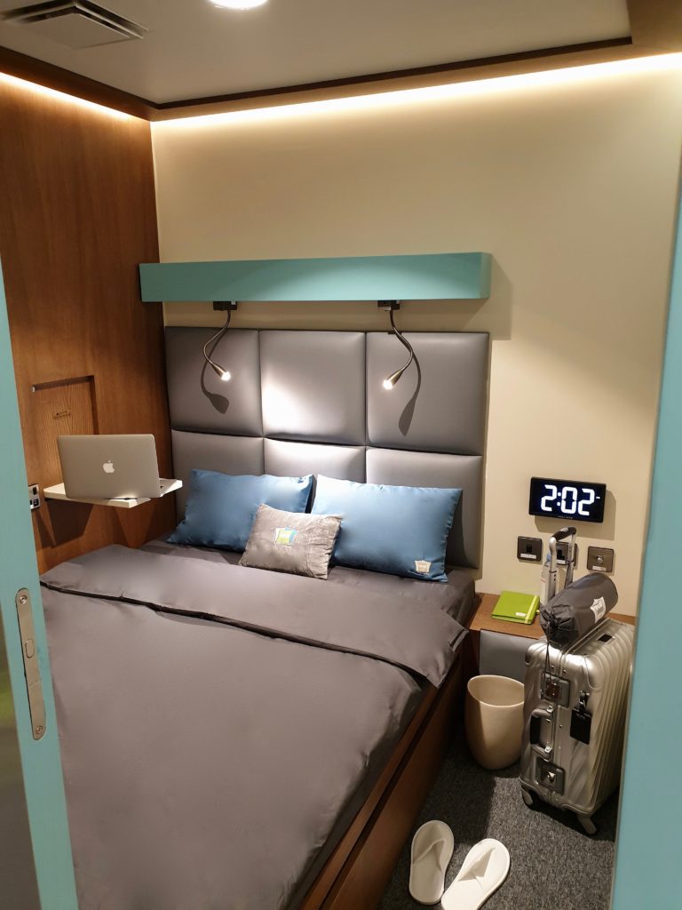 A double bed in one of the sleep cabins, with grey bedding and blue pillows. A pair of slippers are on the floor alongside a passenger bag.