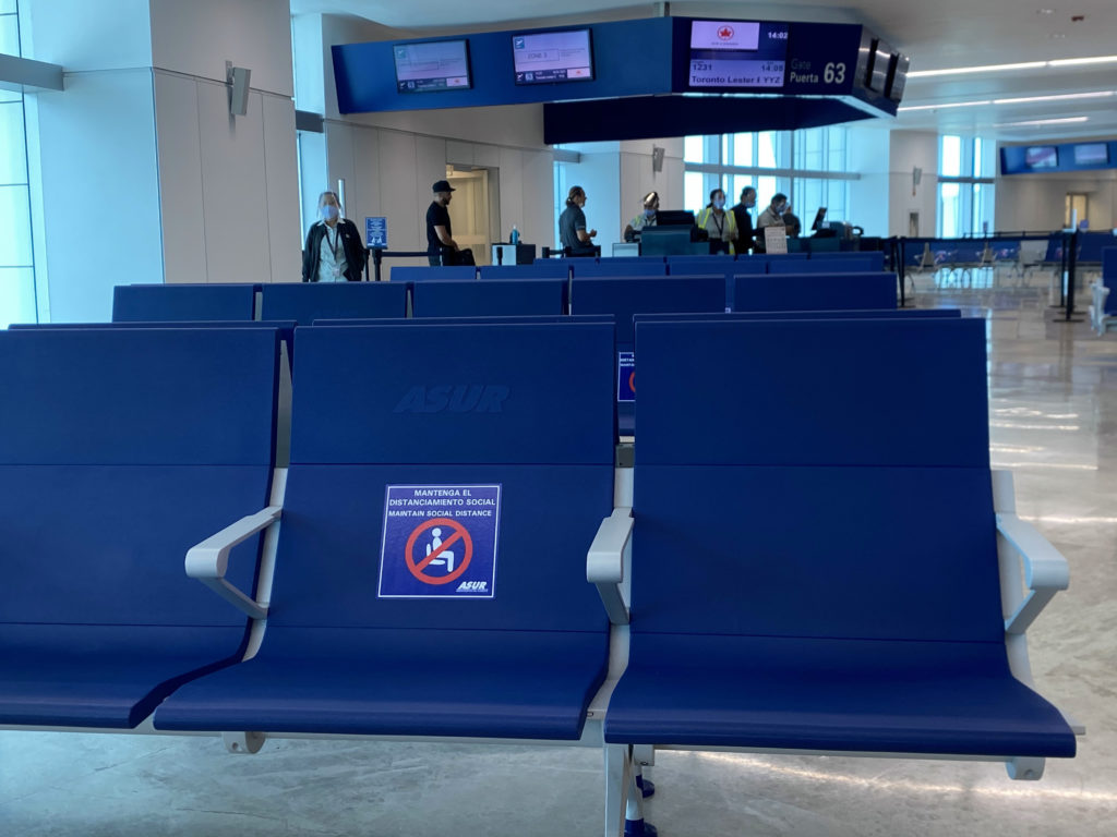 Blue seats at the gate at CUN with screens overhead. A social distancing decal is seen on a seat.