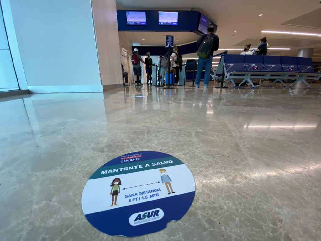 A social distancing marker on the ground at CUN, with screens overhead. Two queues are formed on either side of the agent's desk for boarding