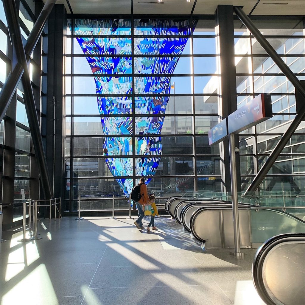 A man and his chid walking towards the escalators at SEA, with a giant blue window art treatment seen in the background