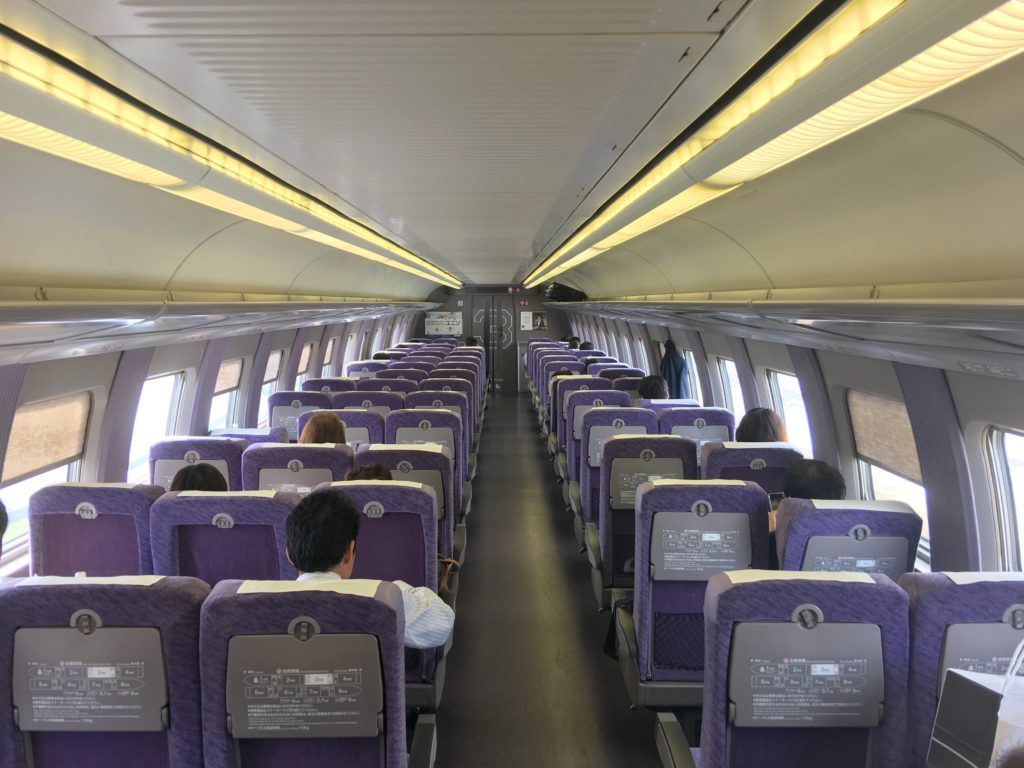 An view from the back of a Shinkansen car showing rows of purple seats, five-abreast (3-2)