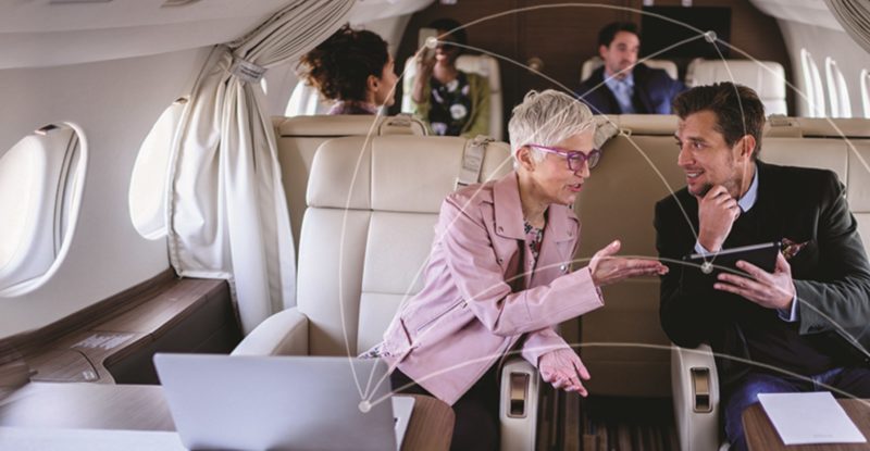 Women in a pick Blazer with shoirt hair talking to a man in a buisness suit on a private Business jet. A few men and women in the background as well.