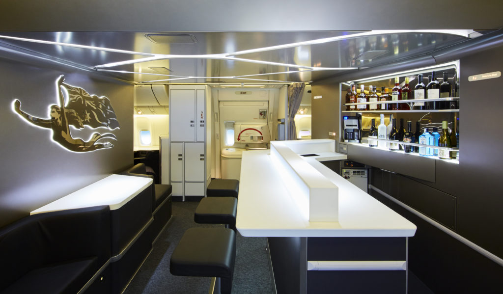 The Virgin Australia bar space, with spectacular lighting and a decorative crest. Two rows of liquor are in view, as well as the long white bar and black stools.