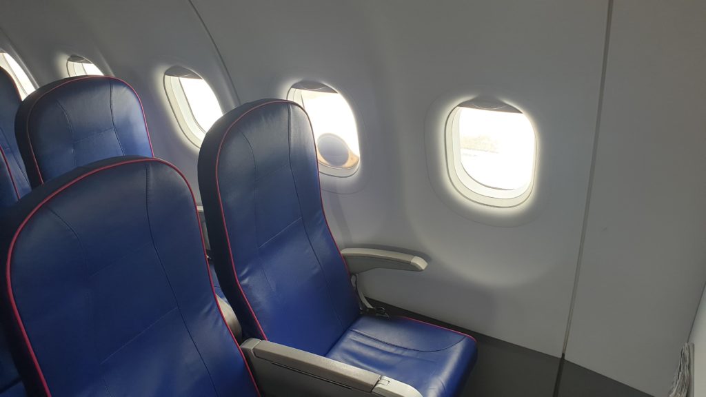 An Iridium antenna attached to an airliner's window, from inside the cabin. The antenna is small and circular. Blue slimline seats are in view.