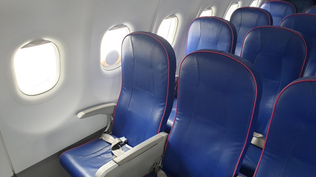 An Iridium antenna attached to an airliner's window, from inside the cabin. The antenna is small and circular. Blue slimline seats are in view. 