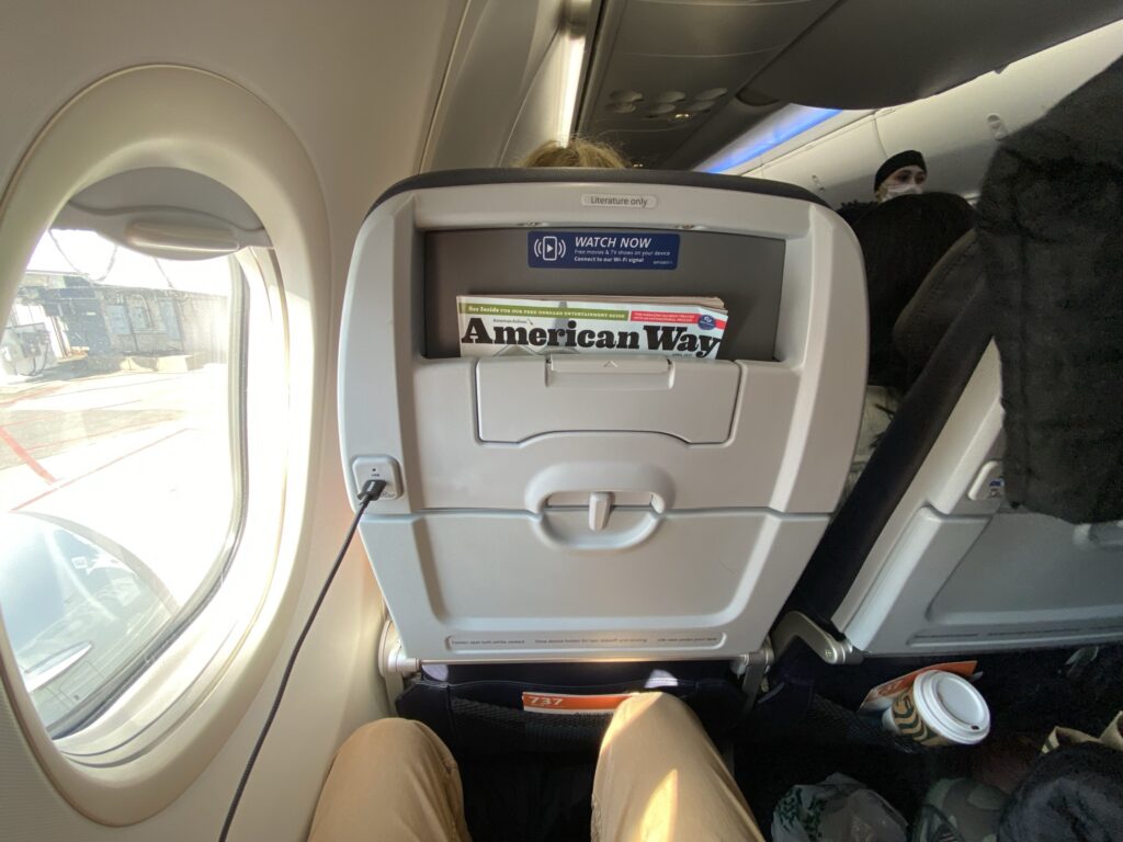 Seatback on the American Airlines Boeing 737-800 in economy class with a ped holder and power port. Man's knees show that there is little space to move.