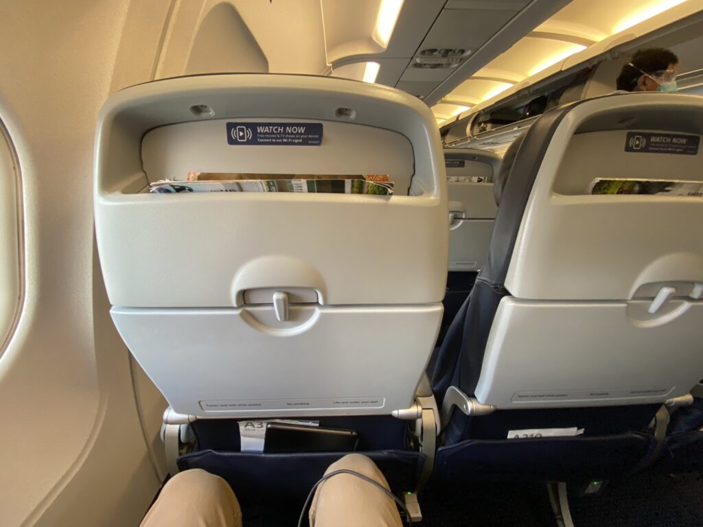 Man's knees showing the space between them and the seat in front of him. Aircraft seat with white tray table and literature pocket. 