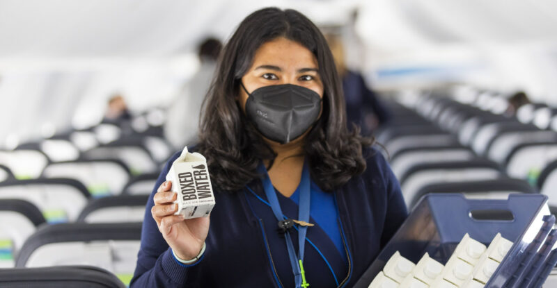 Female Alaska Airlines flight attendant wearing a mask and holding up a boxed water product onboard an aircraft.