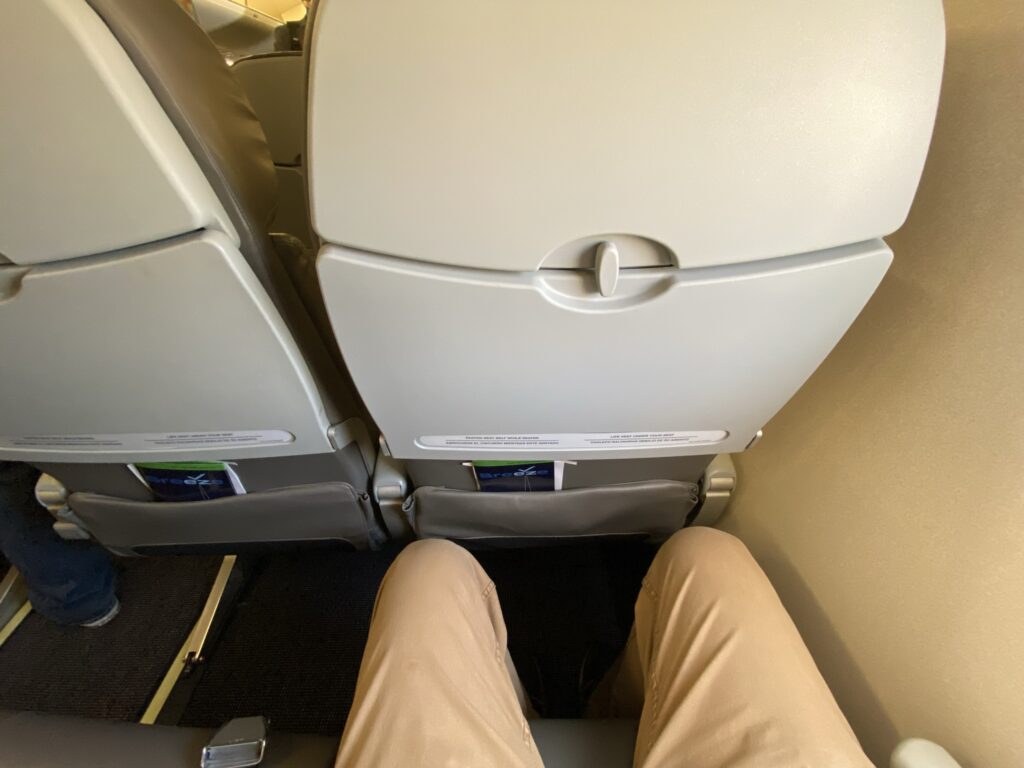 The author shows how much legroom he has in the Breeze economy seat; he has a few inches of knee clearance