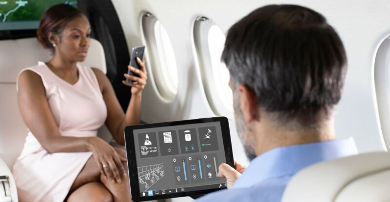 A man and a woman in a private business jet. The woman is holding her mobile device while the man is holding a tablet. The tablet is displaying Collins Aerospace's Venue platform