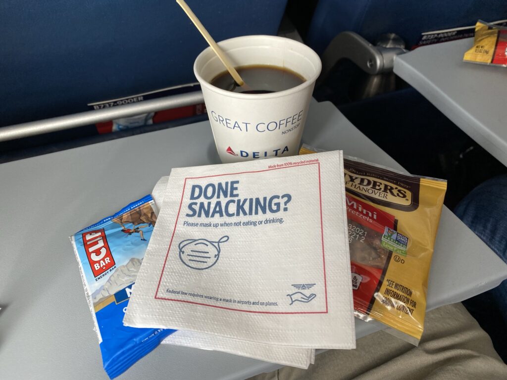 Tray table on the Delta 737 with a coffee, cliff bar and bag of pretzels displayed on it.