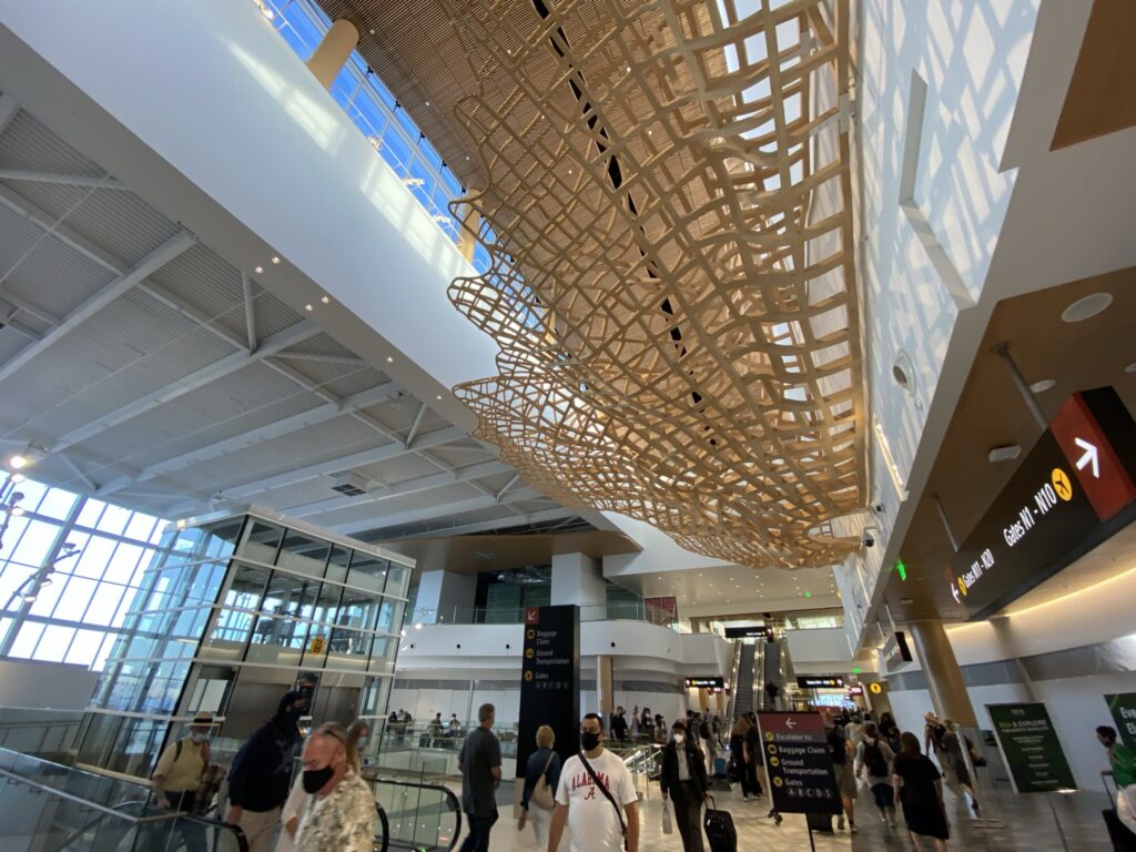 SeaTac's new terminal with many passengers in it. Light is streaming in from windows overhead and on the side of the building, and a unique wooden art installation is in view