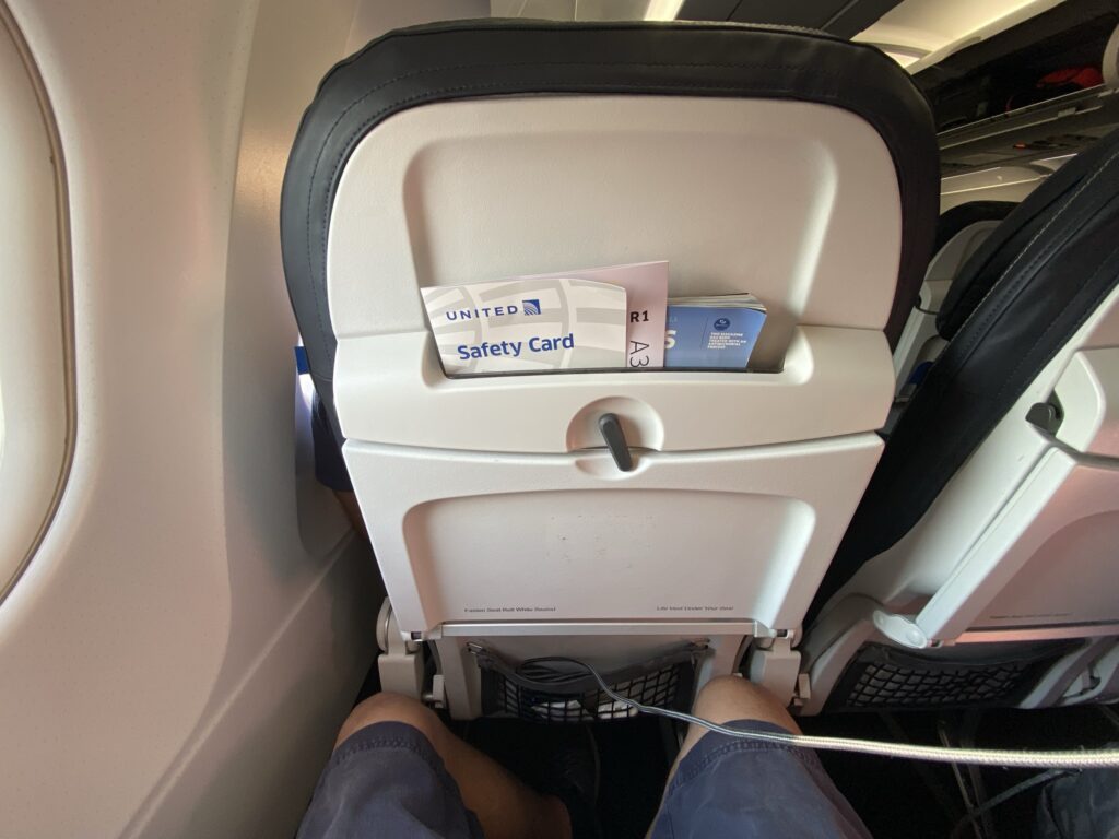 United Airlines A320 Economy class seat. View of a man's knees and the seatback in front of him. There is no device holder, only a high literature pocket and the tray table