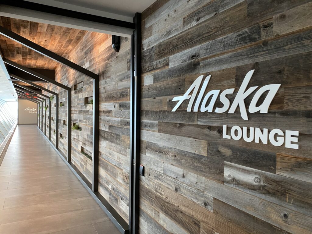 Alaska Airlines' lounge entrance at JFK, with modern wood paneling on the walls and ceiling, and white lettering for "Alaska Lounge"