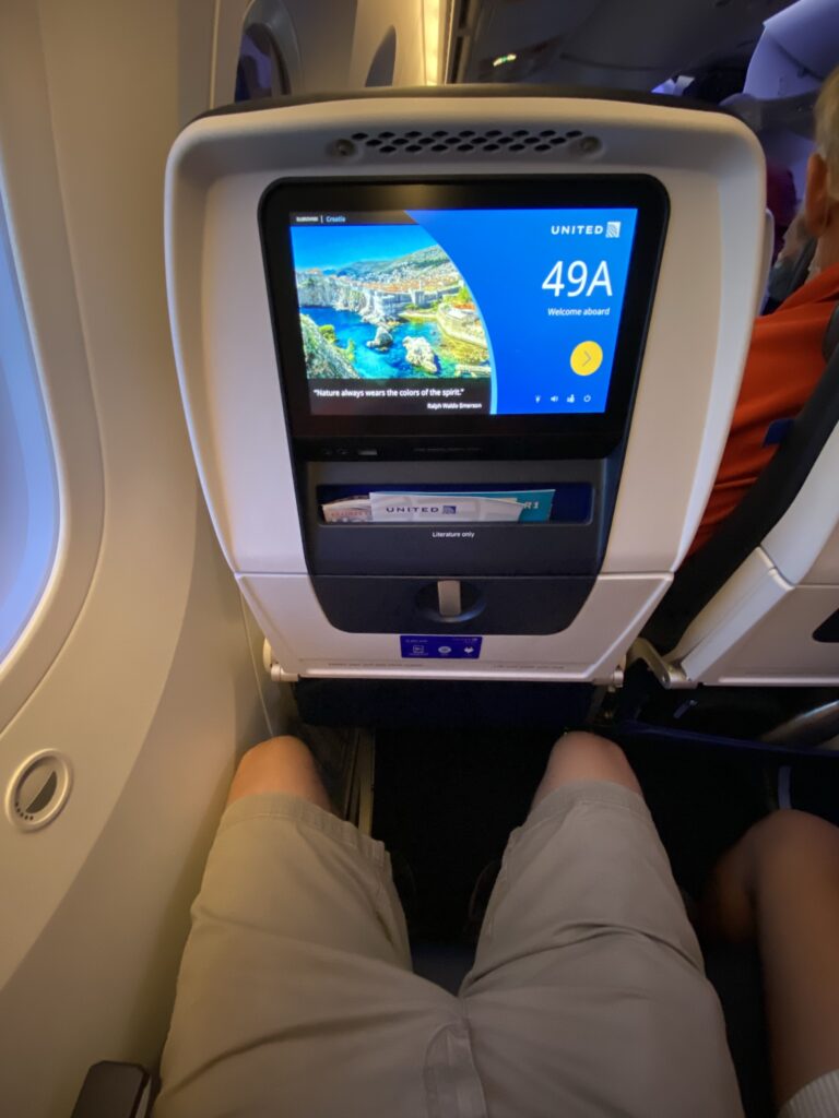 A man's lap and legs are shown sitting in the United Airlines Boeing 787-9 economy class seat. You can also see the embedded IFE in the seatback.