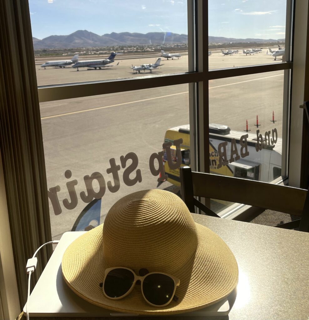 A hat and sunglasses sits on a table. The window view overlooks multiple business jets at Henderson Executive Airport