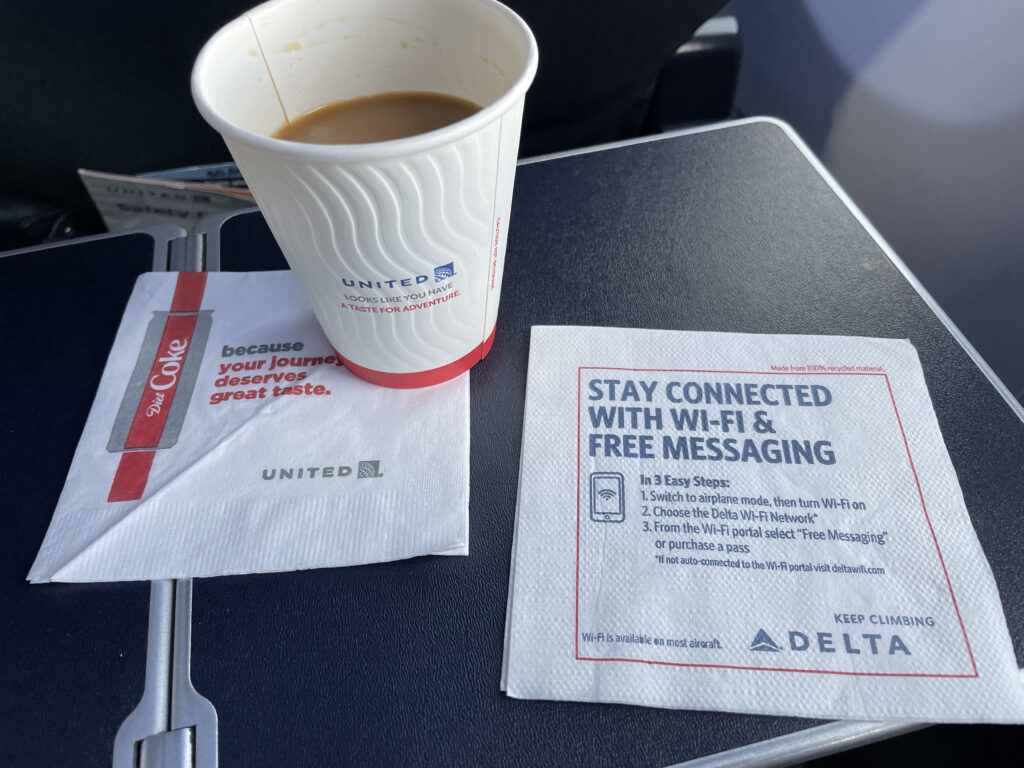 Coffee on an aircraft tray table with both a United napkin and Delta napkin, the latter touting a free text messaging tier of connectivity service