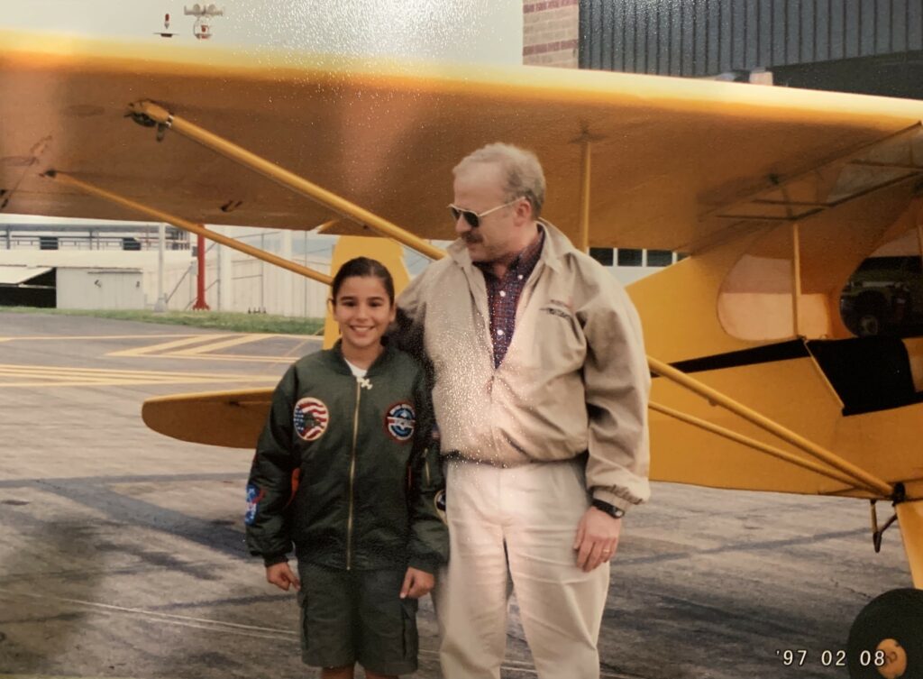 Young Caroline and an elderly male in front of a Yellow Piper Cub aircraft.