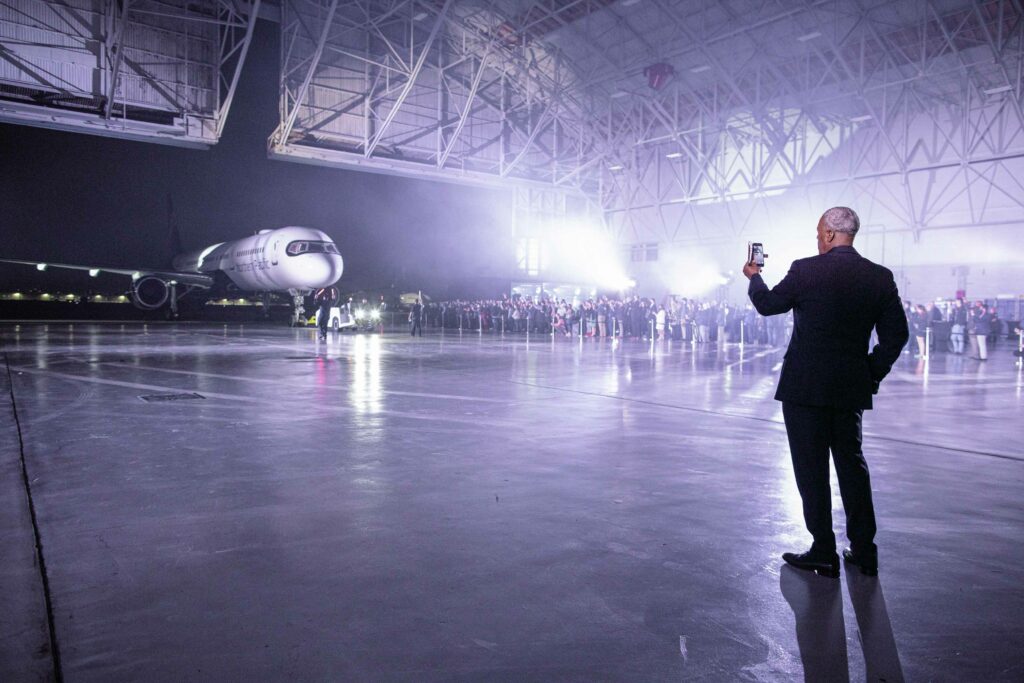 Northern Pacific Airways aircraft reveal. An aircraft sits in the distance of a large hanger as a crowd of people stand around it. A man in a suit stands back to the camera taking a photo with his mobile phone..