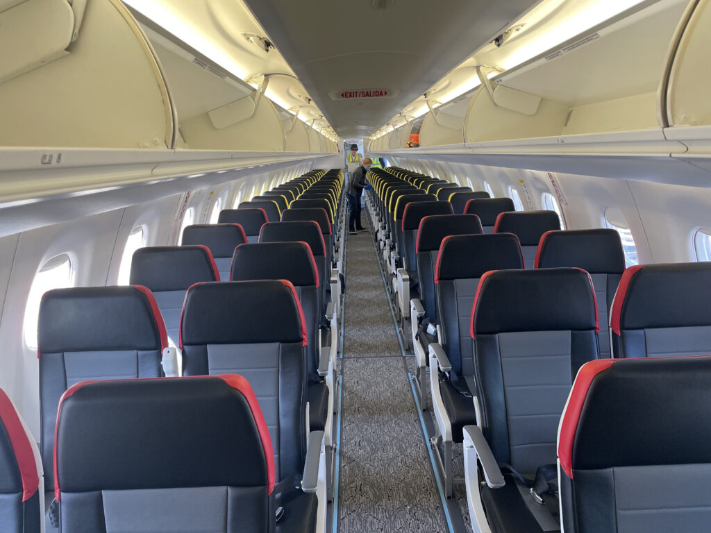 Interior cabin of an aircraft. The seats are all black and grey but the first 5 rows have red trim and the rest have yellow. The seats are in a 2-2 configuration. 