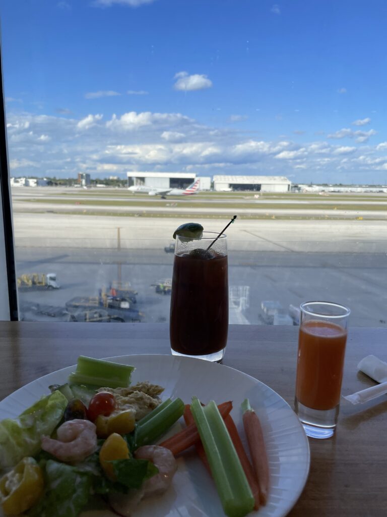 Two drinks and a plate of veggies sit on the lounge table in front of a large window showing the airport runway.