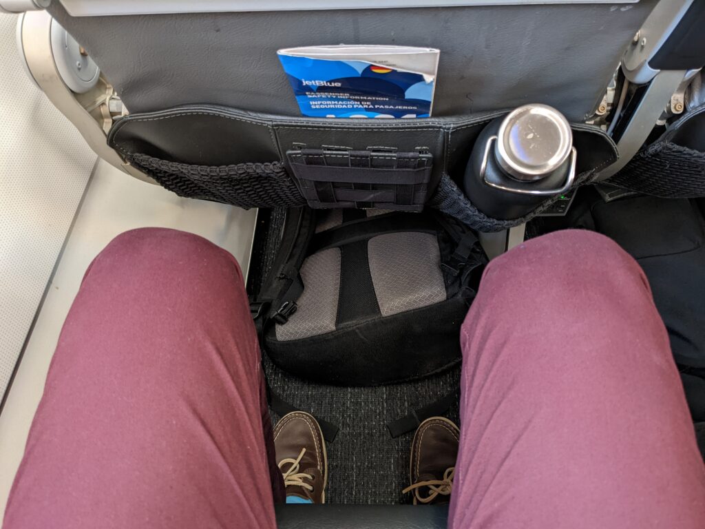 Pictured is a man's legs showing a small gap between him and the aircraft seat in front of him. The pocket on the seatback has a waterbottle placed in it.