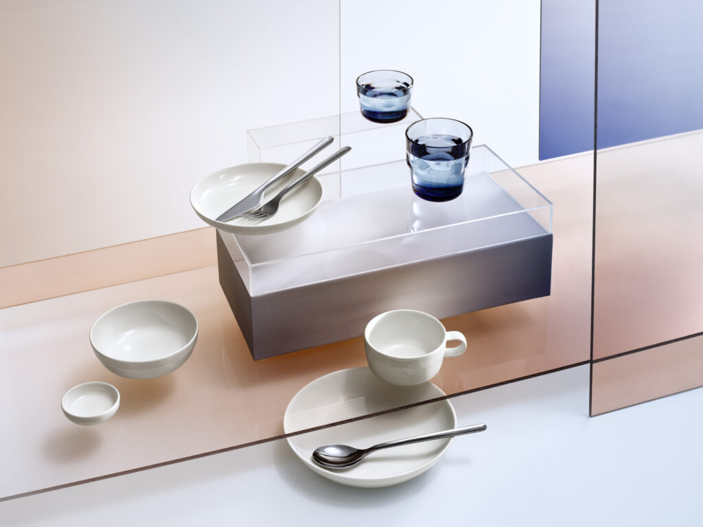 Iittala's new Kuulas tablewear is displayed on a white back drop and wooden table runner. Two glasses with blue details sit beside a bowl with utensils in it. More dishes sit below it including a tea cup.