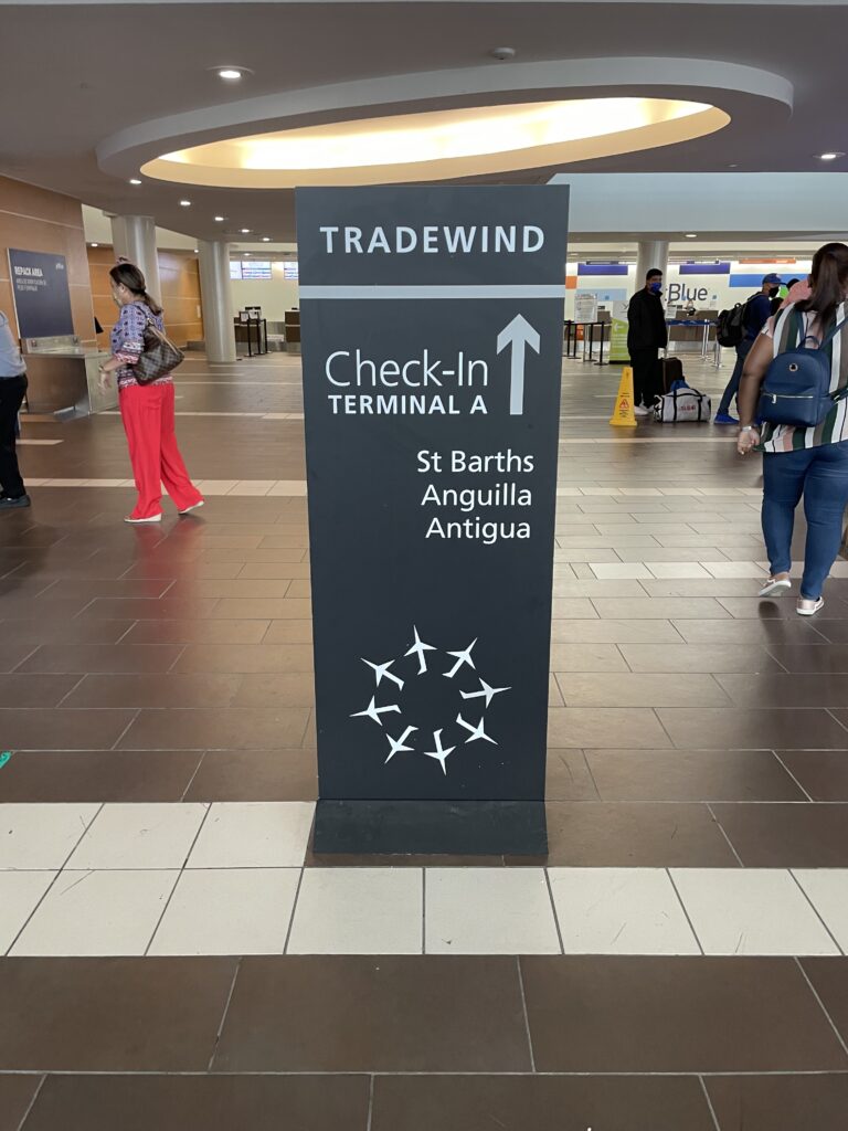 Tradewind check-in sign is displayed in Luis Munoz Marín International Airport. Passengers walk about the airport.