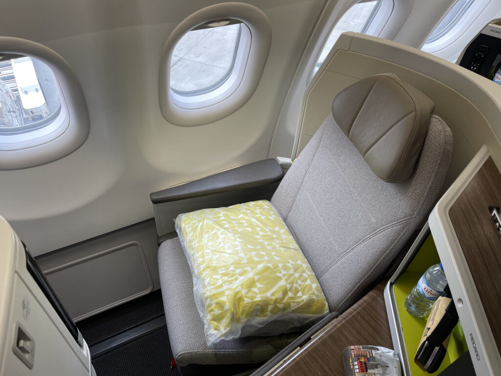 A window seat on TAP A330-900neo business class. The seat is grey with a yellow pillow resting on top.