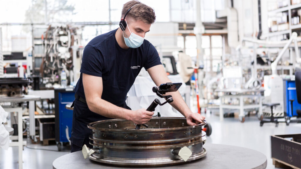 A masked LHT employee holding a camera towards an aircraft engine part for the 5G campus network