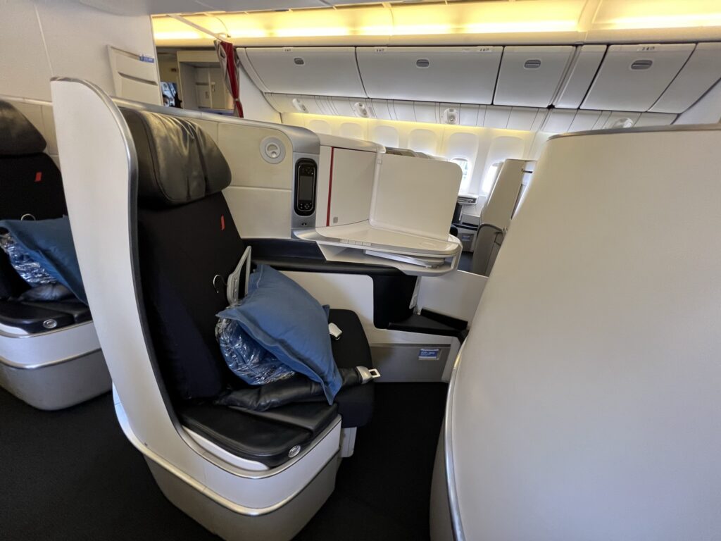 Air France Boeing 777-300ER business class seat. The seat has a pillow, blanket and hanger sitting on it. The seat is dark blue with grey thermoplastics wrapping around it.