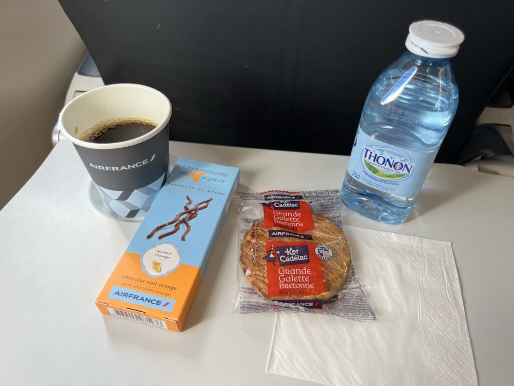 A cup of coffee, bottle of water, packaged pastry and chocolate sticks are sitting on an aircraft tray table.