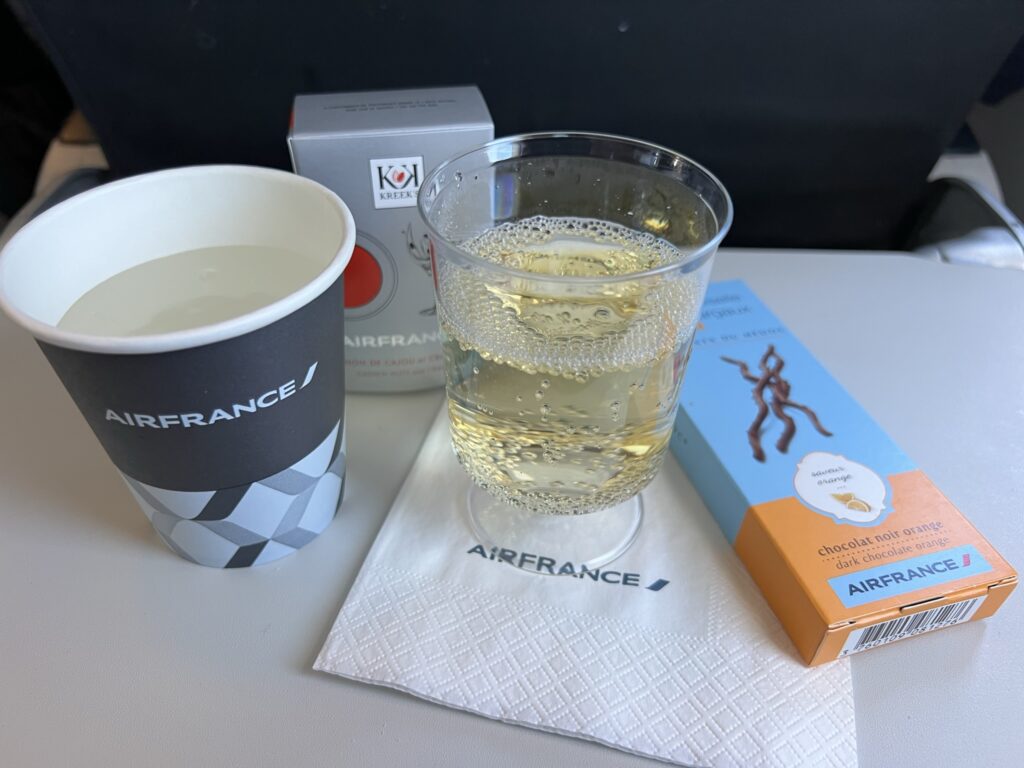 A cup water, glass of champagne and a box of chocolates sits on an Air France tray table.