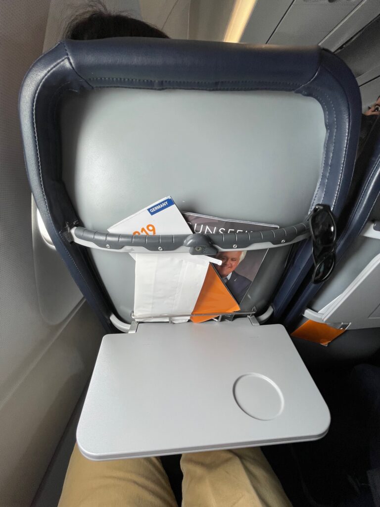 Tray table is flipped open on the aircraft seat and a literature pocket is visible and holding a safety card and inflight magazine.